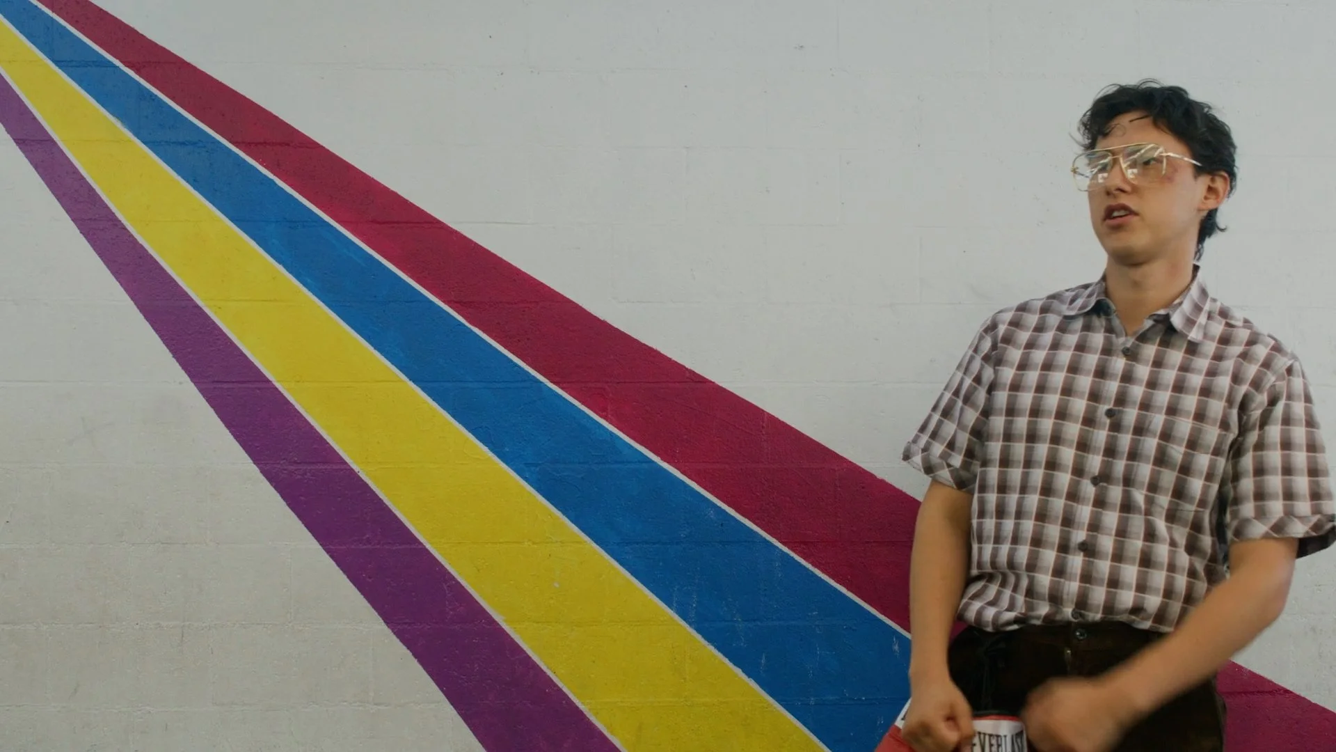 A young man with curly black hair, wearing glasses, a brown and white checkered shirt, standing in front of a white wall with multicolored diagonal stripes in purple, yellow, blue, and red.