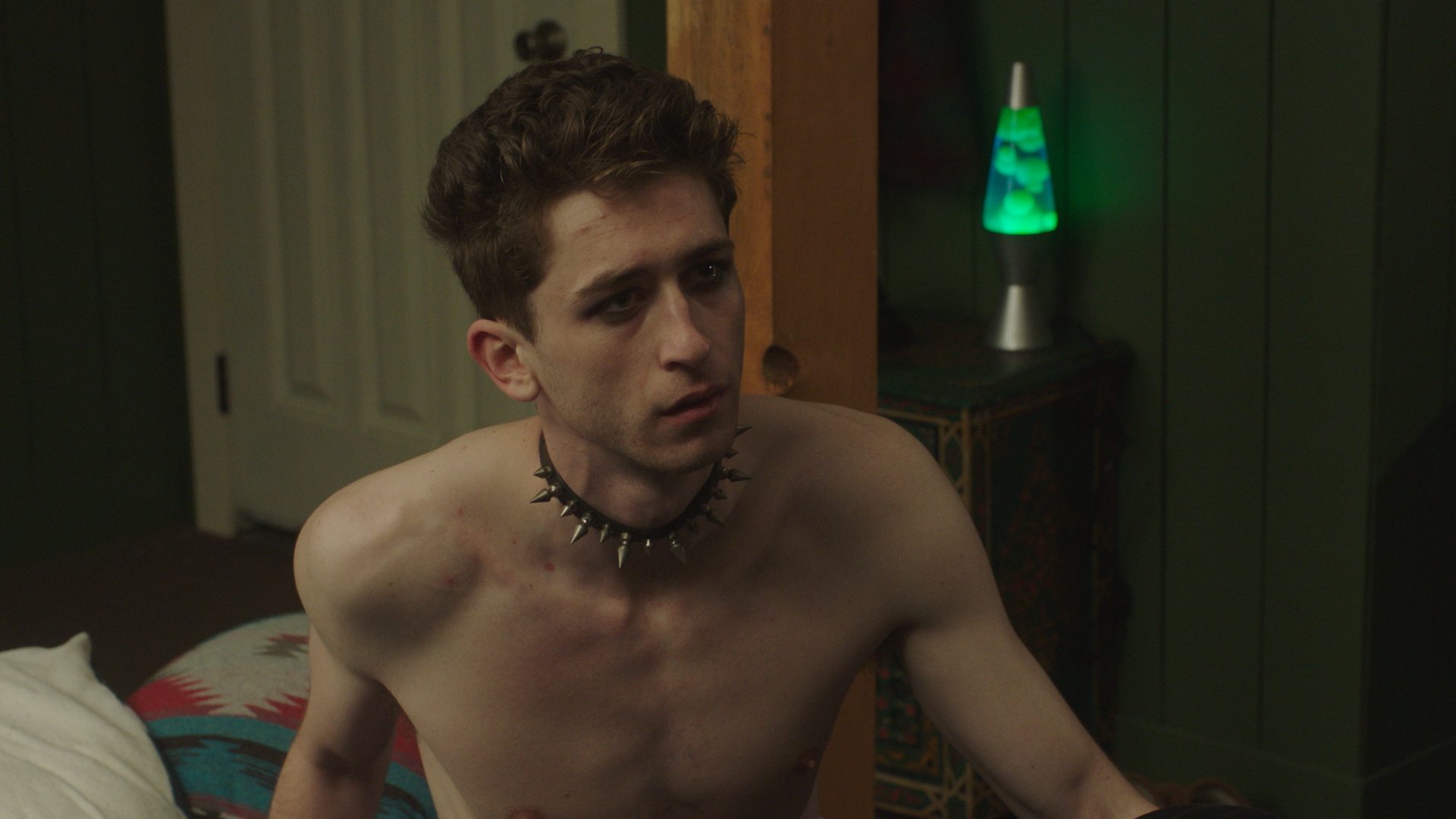 Young man with dark hair, wearing a spiked choker, shirtless, sitting on a bed in a dimly lit room with green walls and a lava lamp in the background.