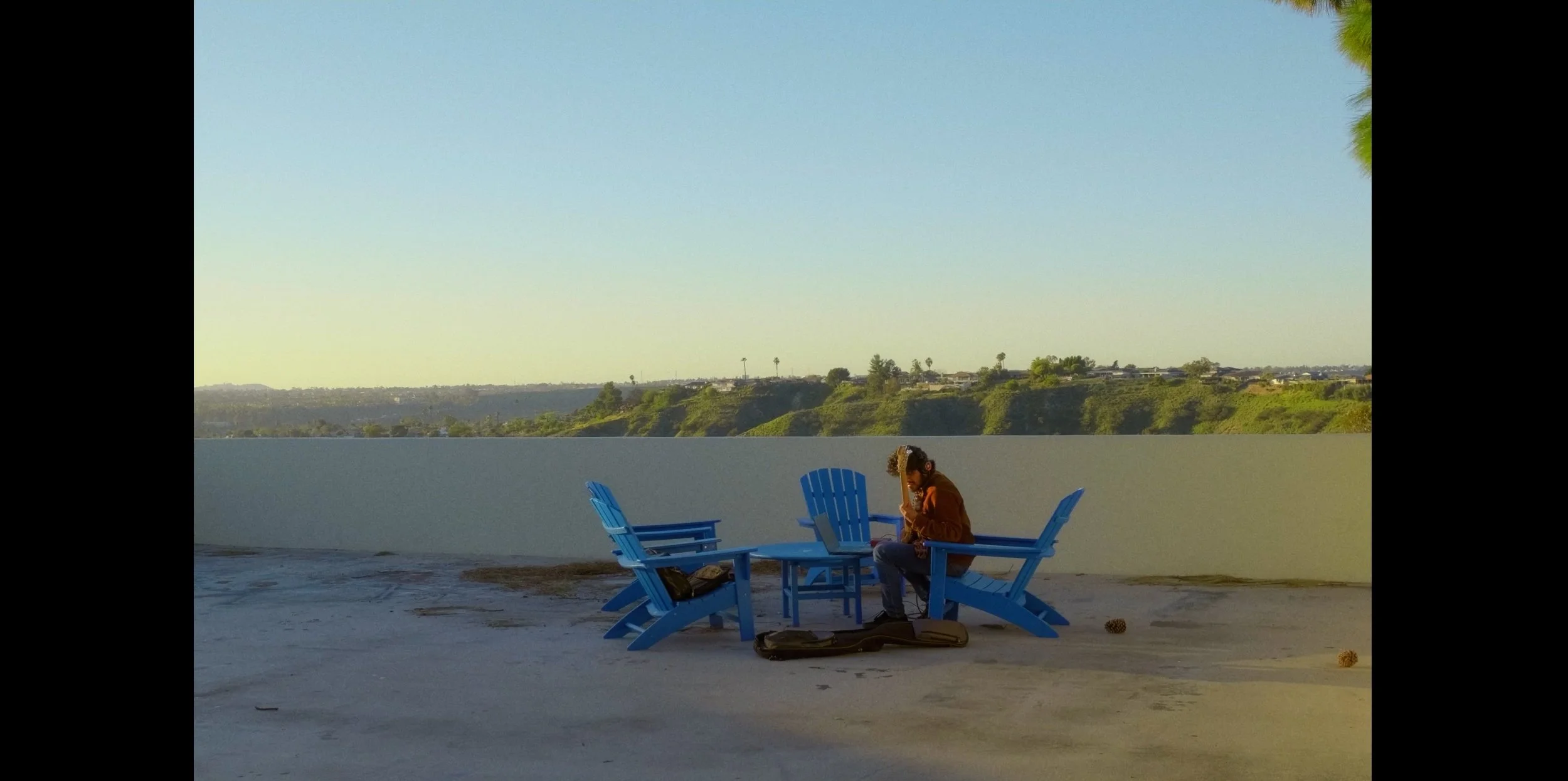 Person sitting on a blue Adirondack chair on a rooftop, with a guitar case and two pine cones nearby, overlooking a landscape with hills and houses in the distance under a clear sky.