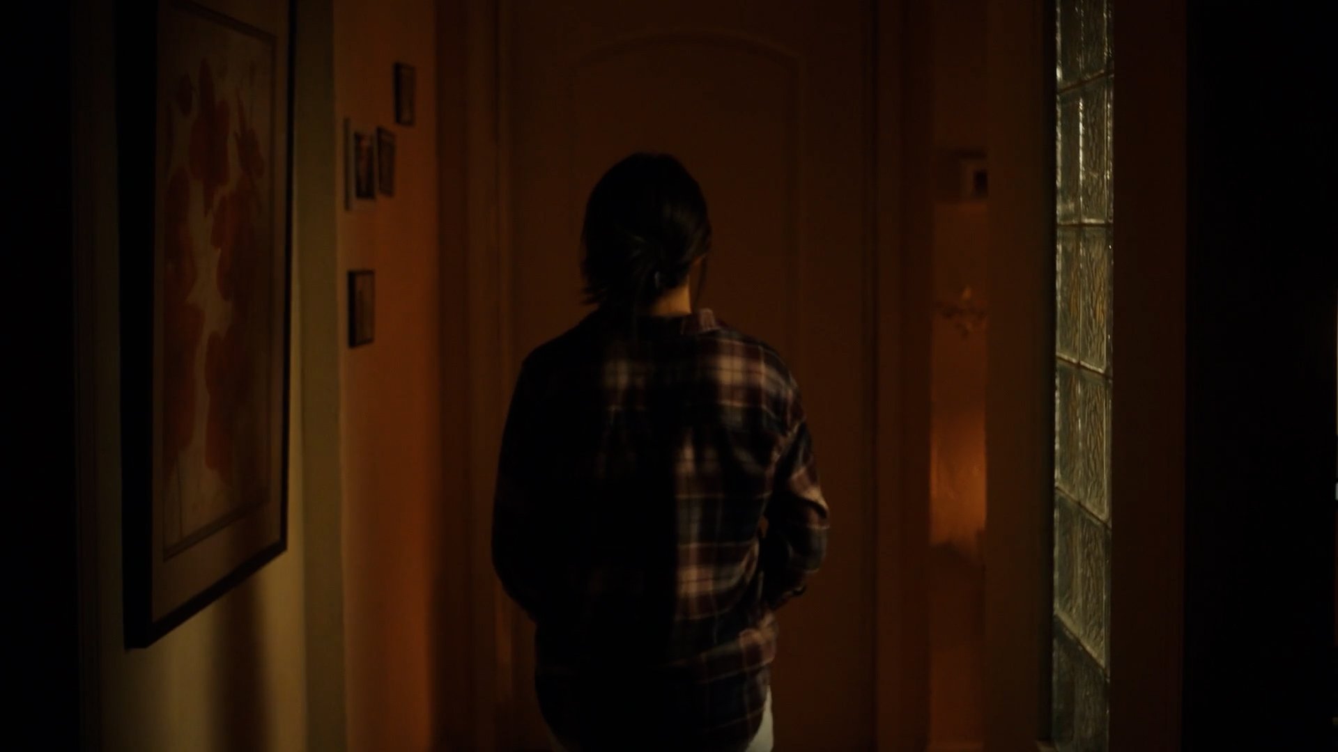 Person with dark hair wearing a plaid shirt standing in a dimly lit hallway, looking towards a door.