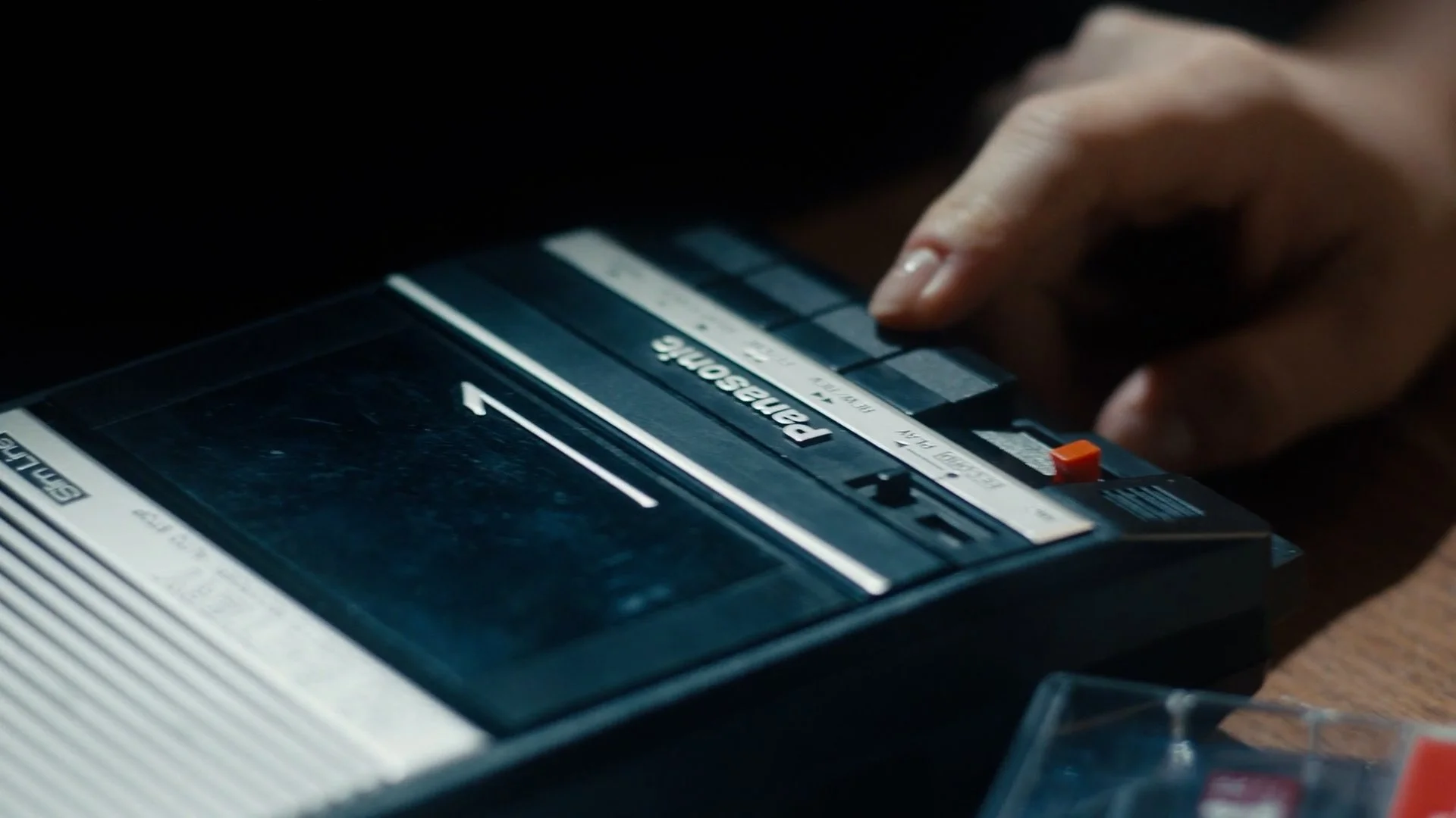 A hand reaching to push a button on a vintage Panasonica tape recorder. The tape recorder is black with white and gray details and is placed on a wooden surface.