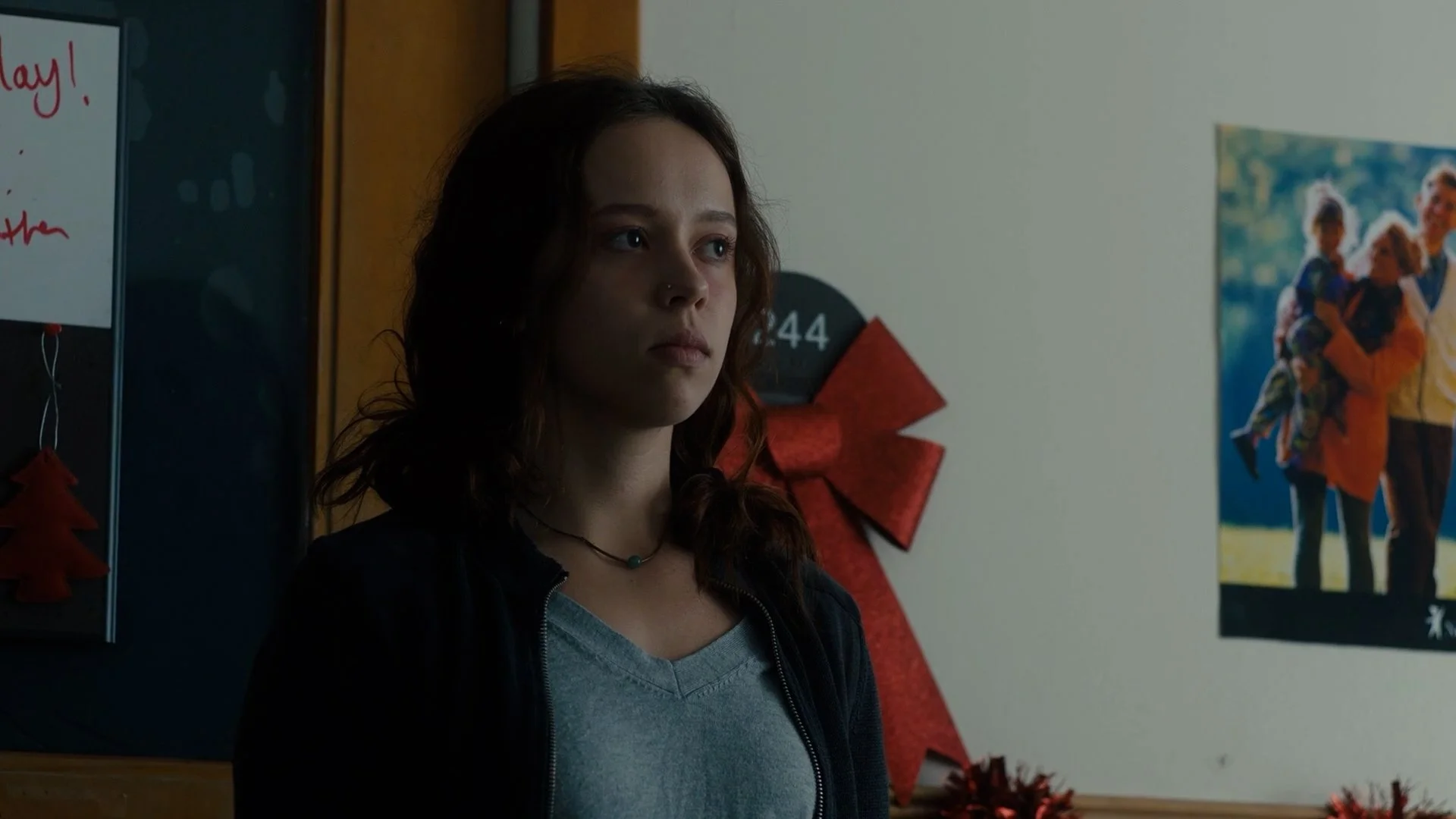 Young woman with wavy brown hair, wearing a gray shirt and a black jacket, standing indoors near Christmas decorations, including a red bow and a holiday poster with two children and an adult.