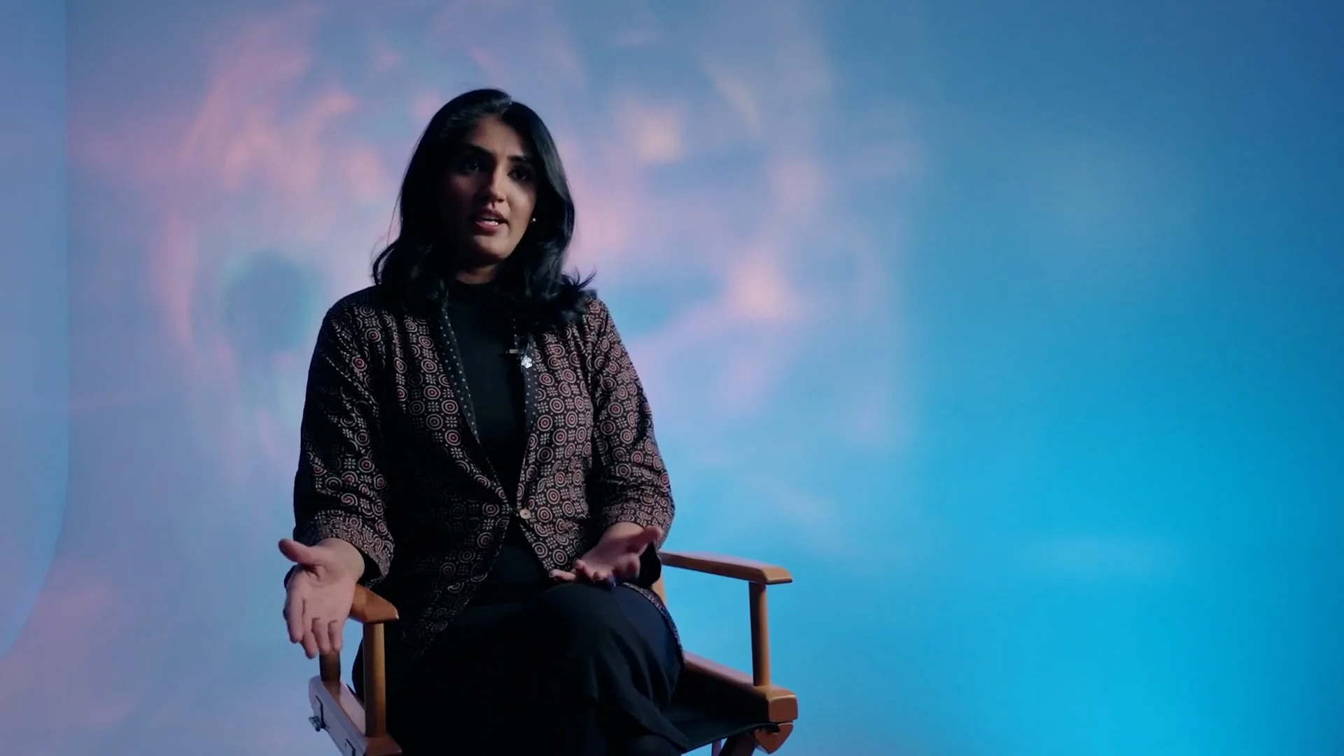 A woman with dark hair seated on a director's chair, speaking against a blue sky backdrop with pink clouds.