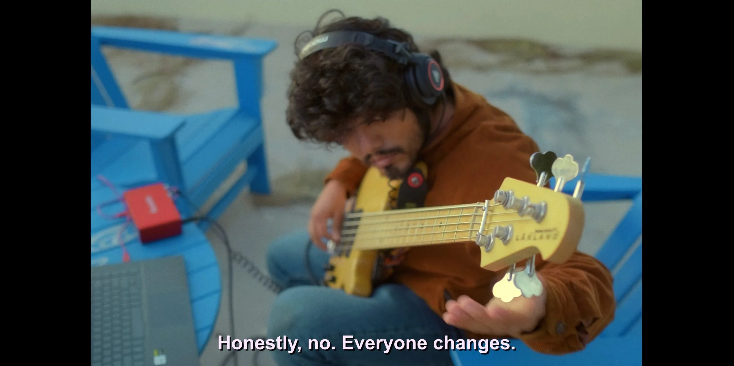 A man sitting on a blue chair playing a yellow bass guitar with a capo. He is wearing headphones and a brown shirt. There is a small red speaker and some electronic devices on the blue table next to him. Subtitles at the bottom say, 'Honestly, no. Ev