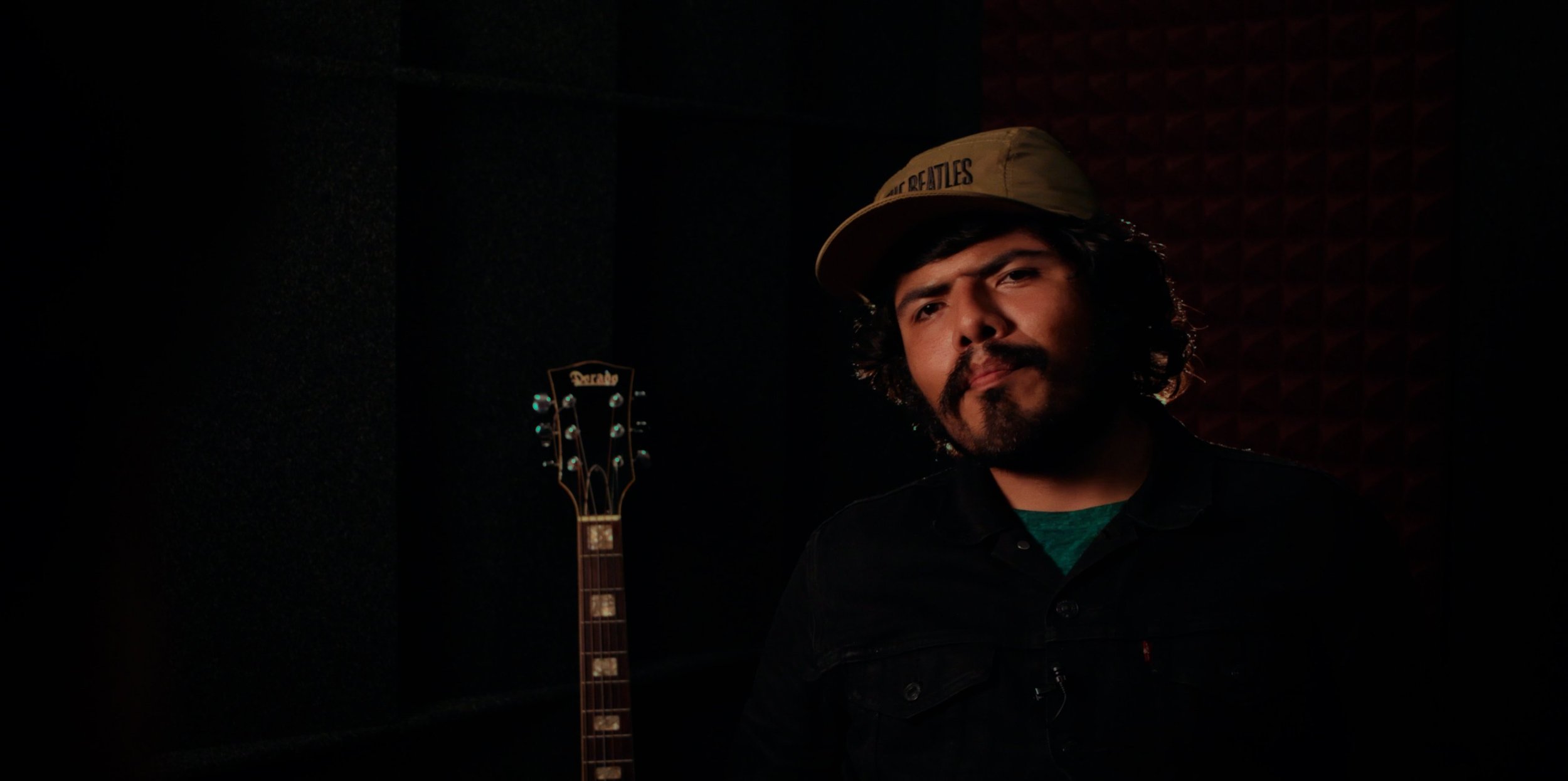 A man with curly dark hair, beard, and mustache wearing a beige cap and black jacket, seated next to a guitar in a recording studio with soundproofing panels.