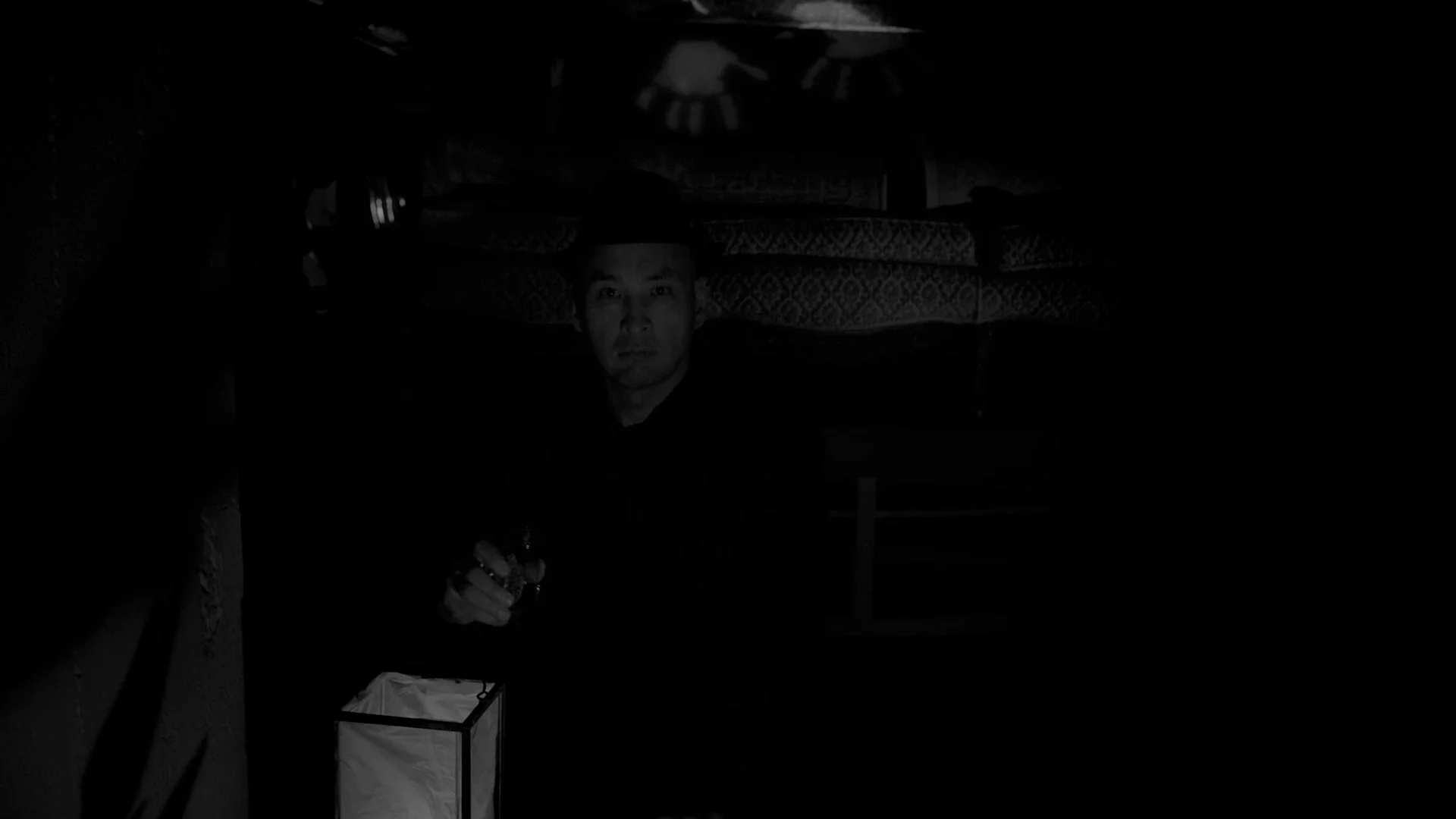 A person with a hat sitting in a dark room, with shelves behind them and a lamp on the table in front.