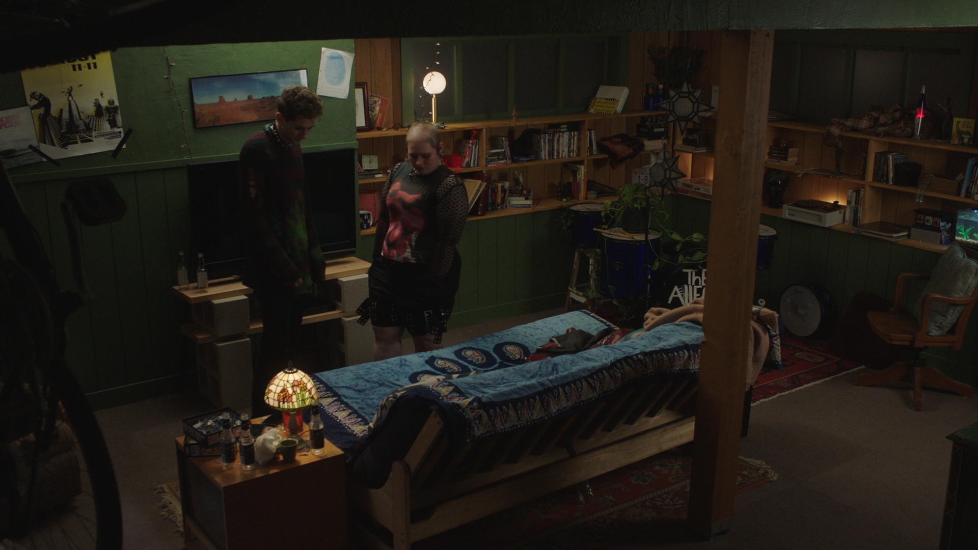 Darkly lit room with two women standing near a bed covered with a blanket, surrounded by shelves filled with books and DVDs, with a small table holding bottles and a lamp, and a chair in the corner.