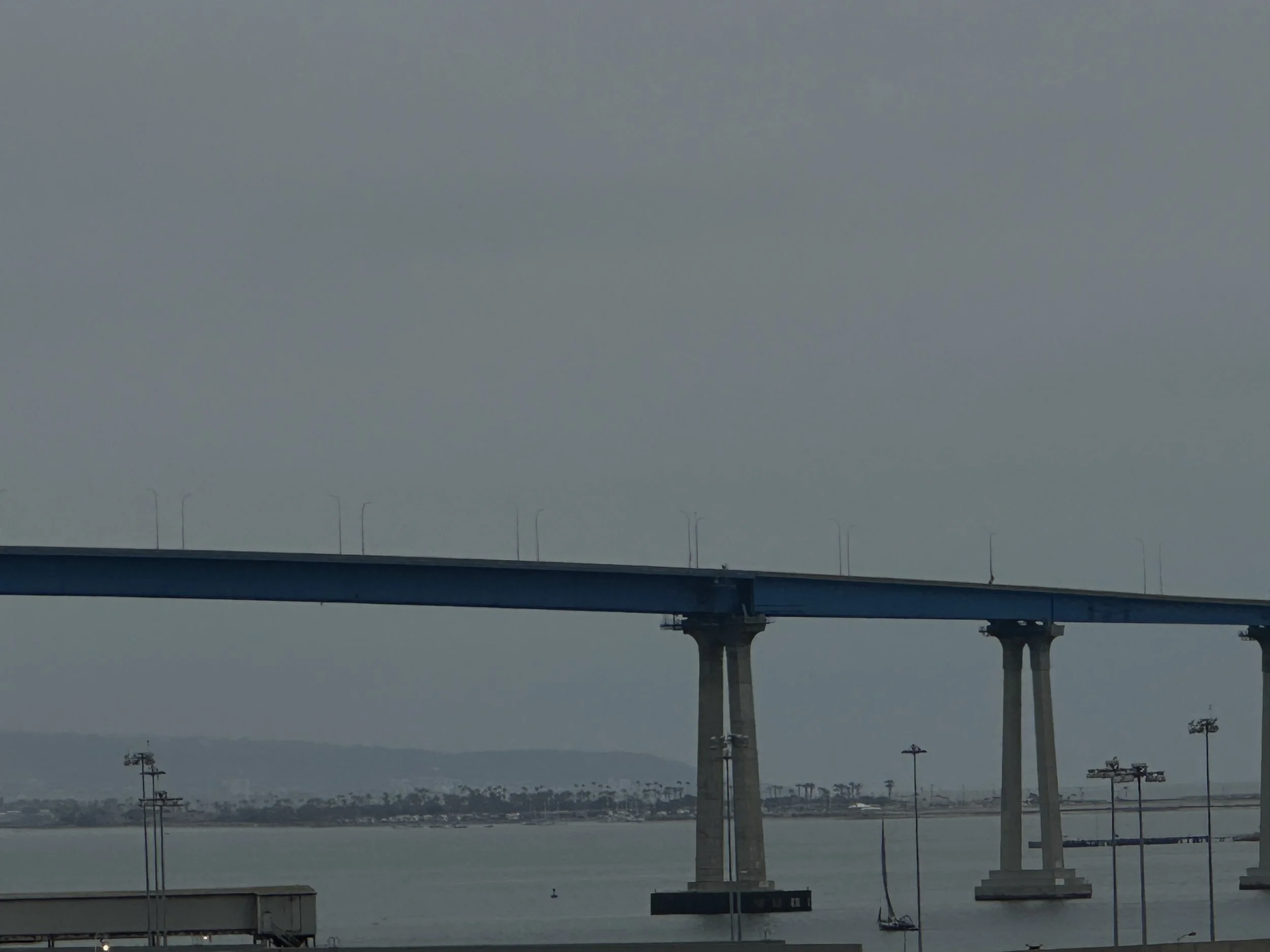 A large bridge with concrete supports stretches over a body of water, with land and hills visible in the distance under a cloudy sky.