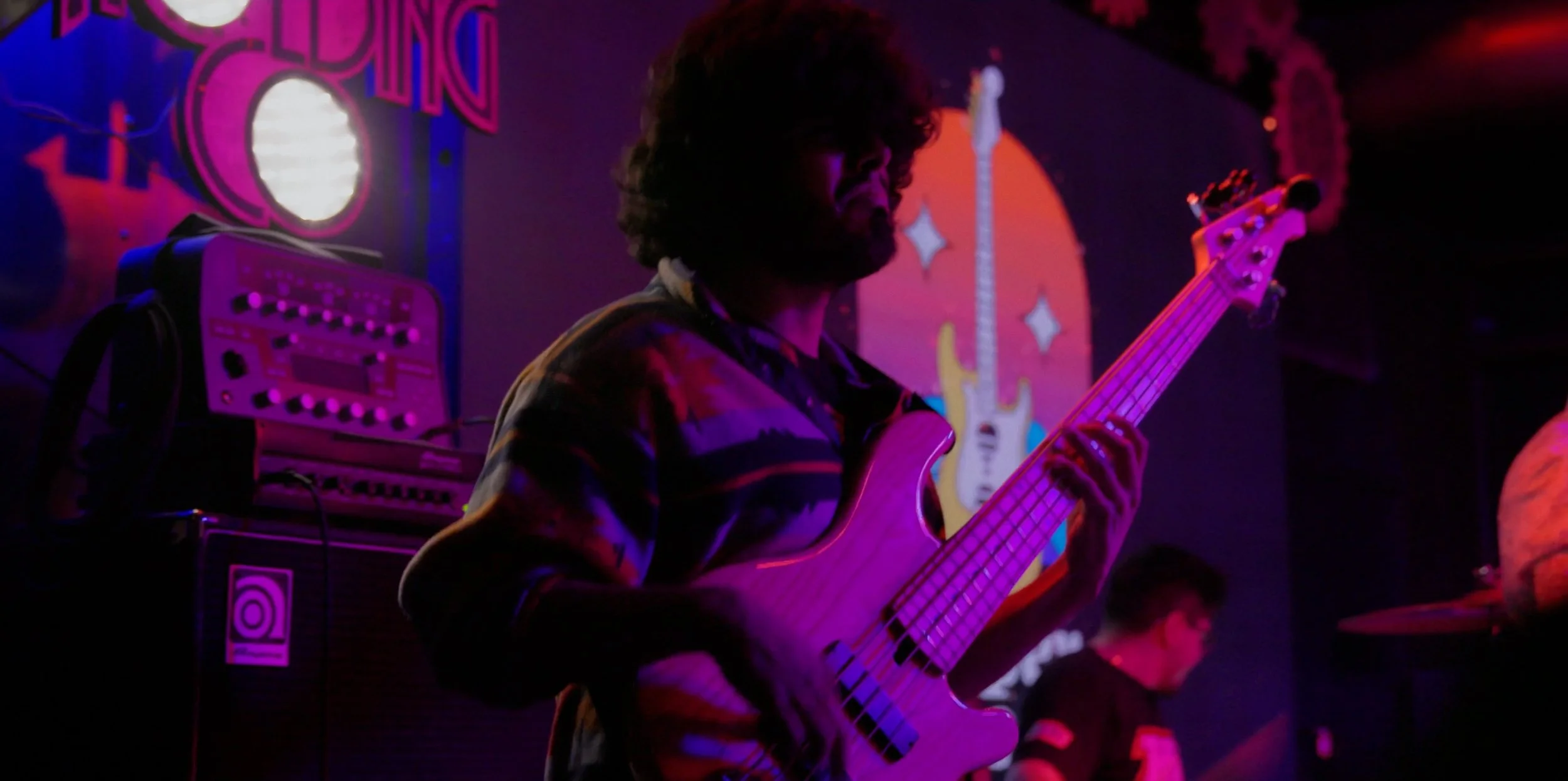 Musician playing a bass guitar on stage illuminated with purple and pink lighting, with musical equipment and a colorful backdrop in the background.