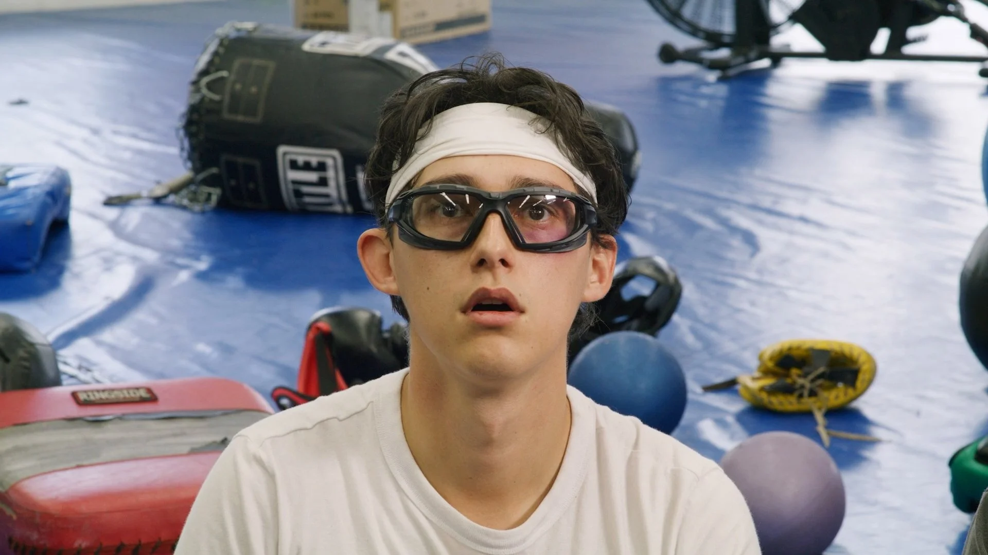 A young man with curly dark hair wearing sports goggles, a white headband, and a white shirt, sitting on the floor of a gym or training area filled with boxing and sports equipment.