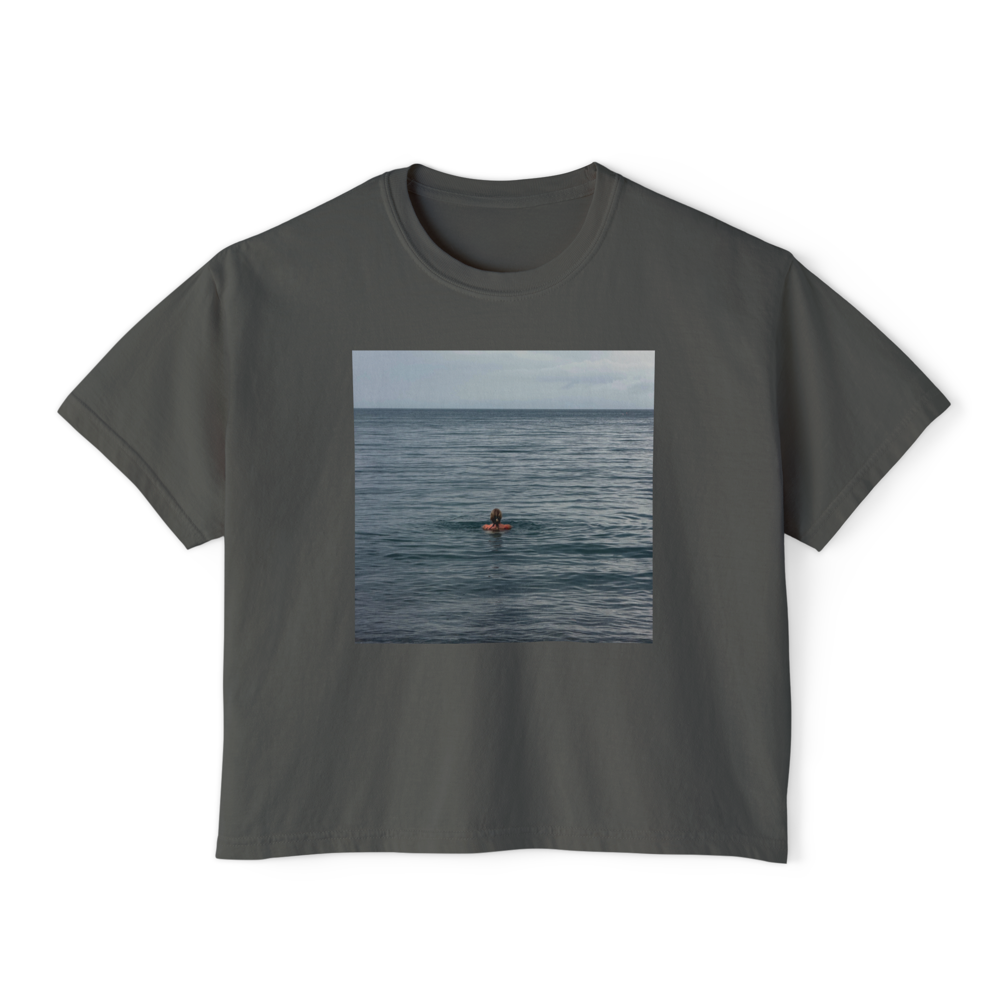 A white T-shirt with a rectangular photo of a person floating on their back in the ocean, facing away from the camera, with water and sky in the background, displayed on a black background.