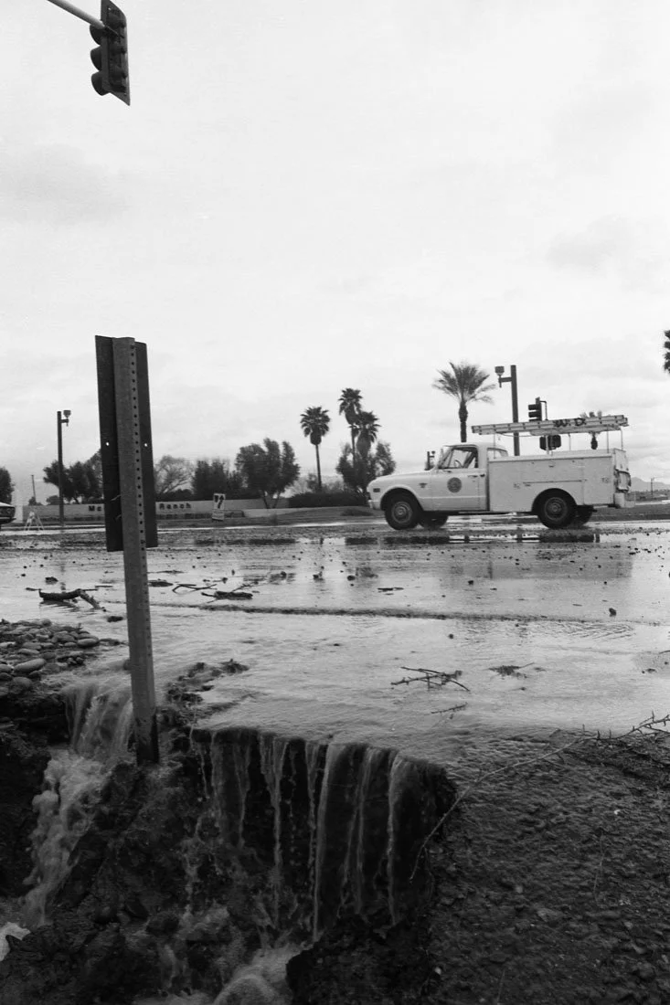 A flooded street with water flowing over the curb, a white utility vehicle on the road, and palm trees in the background under a cloudy sky.