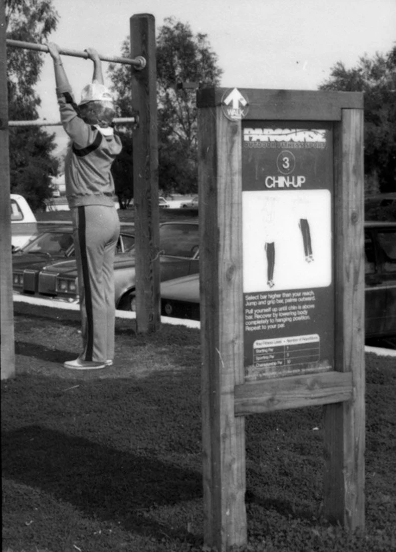 A person wearing a helmet and a sports outfit is hanging from a horizontal bar at a playground. There is a signboard nearby with instructions for an activity.