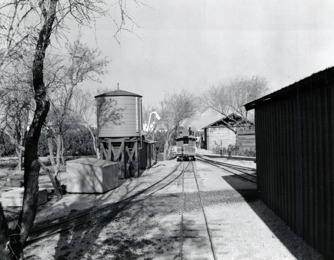 A black and white photo of a train approaching a small train station with tracks, trees, and small buildings around.