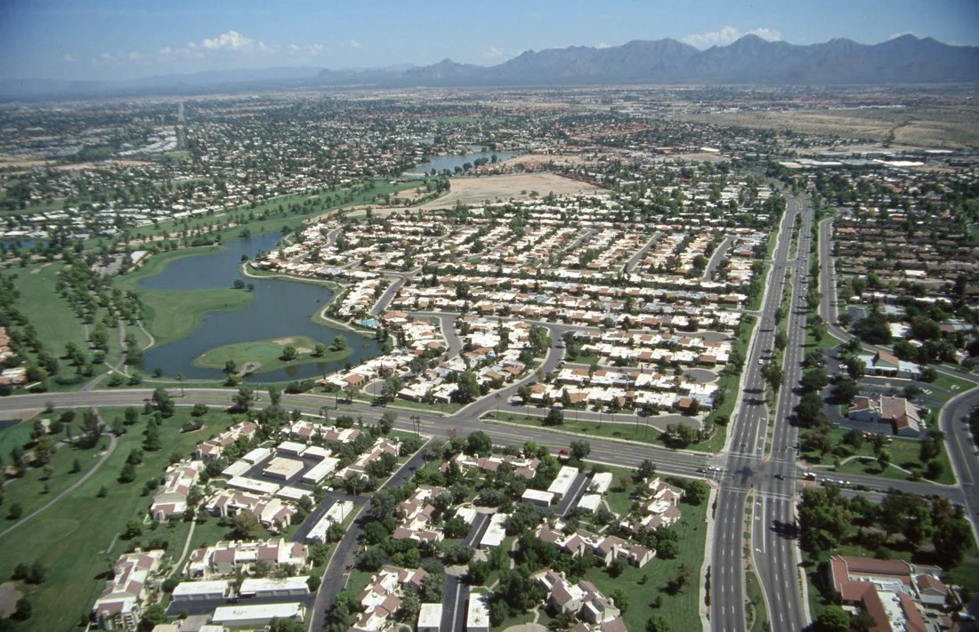 Aerial view of a suburban neighborhood with lakes, roads, and green spaces, mountains in the background.