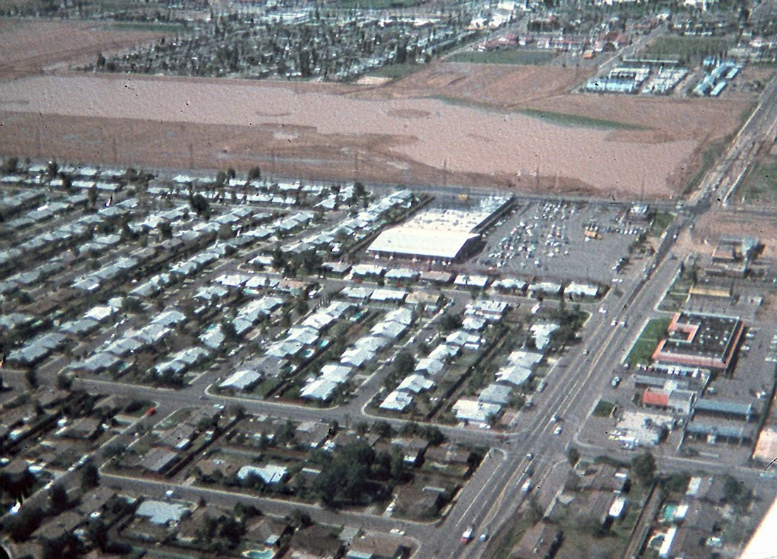 Aerial view of a suburban area with residential houses, commercial buildings, parking lots, and a main road, with open fields in the background.