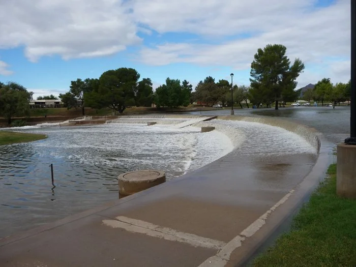 A river overflowing onto a paved walkway in a park with trees, grass, and cloudy sky.