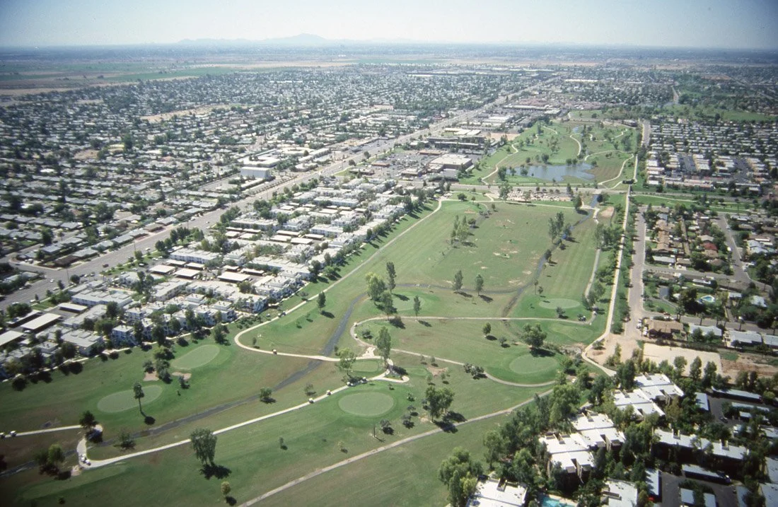 Aerial view of a city with a large park featuring golf course greens, walking paths, lakes, and trees.