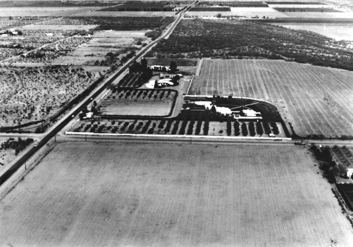 Aerial black and white photograph of a farm estate with multiple buildings, well-maintained gardens, and fields in the background, divided by roads.