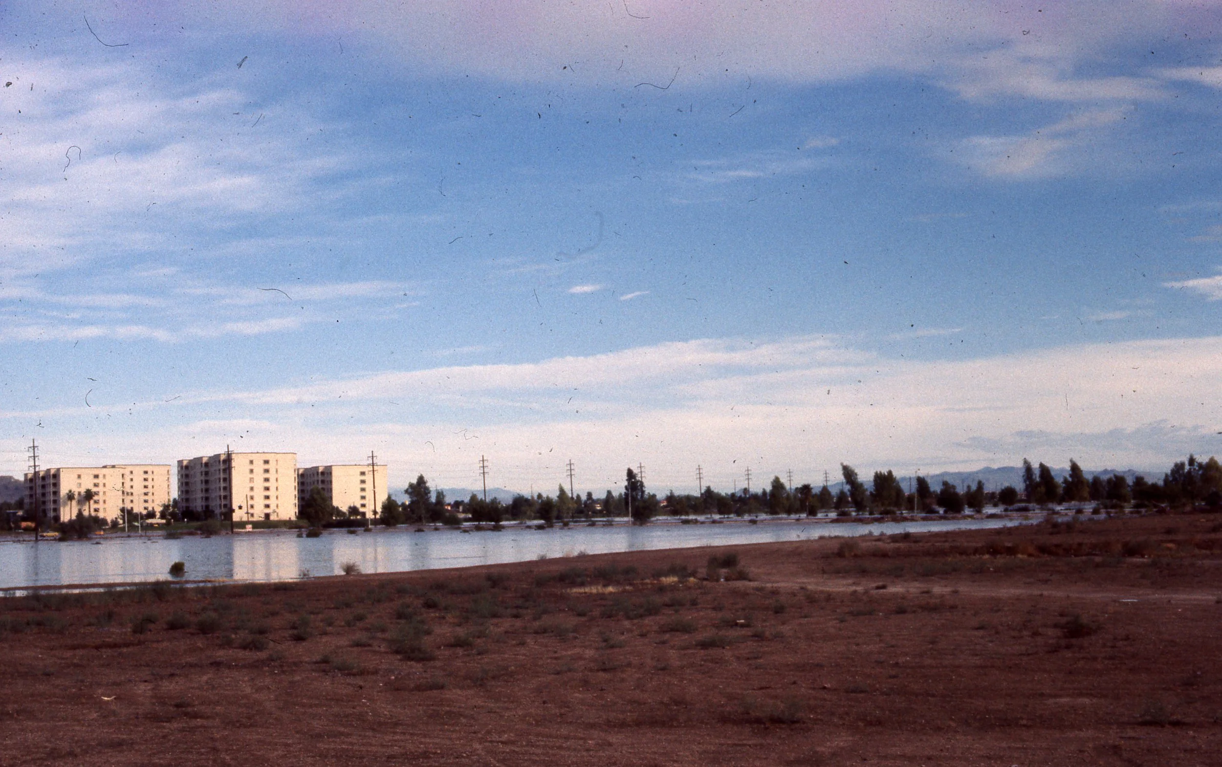 A landscape scene with a body of water in the foreground, modern apartment buildings in the middle distance, numerous utility poles and power lines, trees, and mountains in the background under a partly cloudy sky.