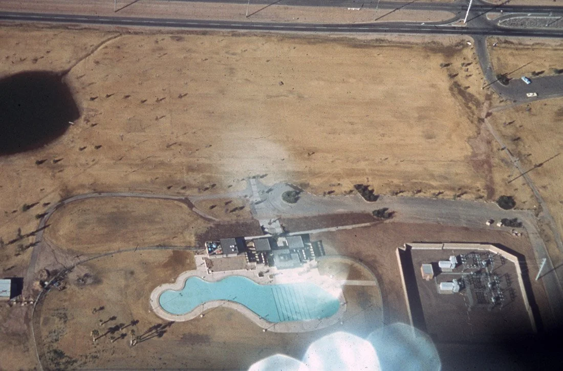 Aerial view of a residential area with a small community pool, surrounding buildings, and streets in a desert landscape.