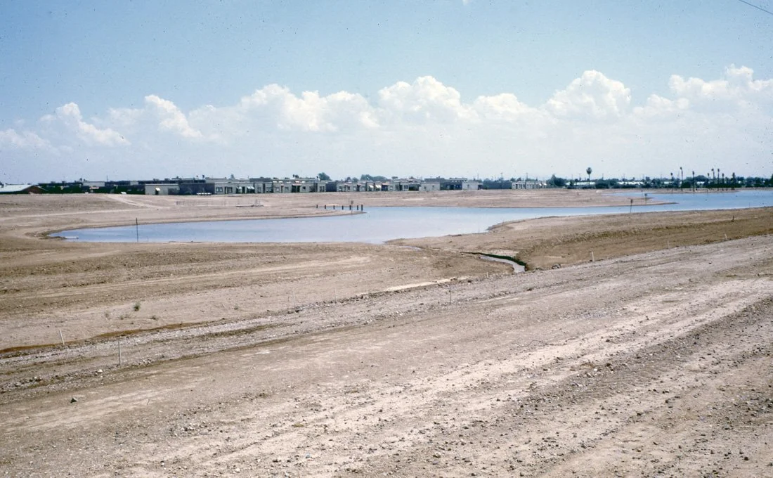 A barren construction site with a small water reservoir and a row of modern apartment buildings in the background under a partly cloudy sky.