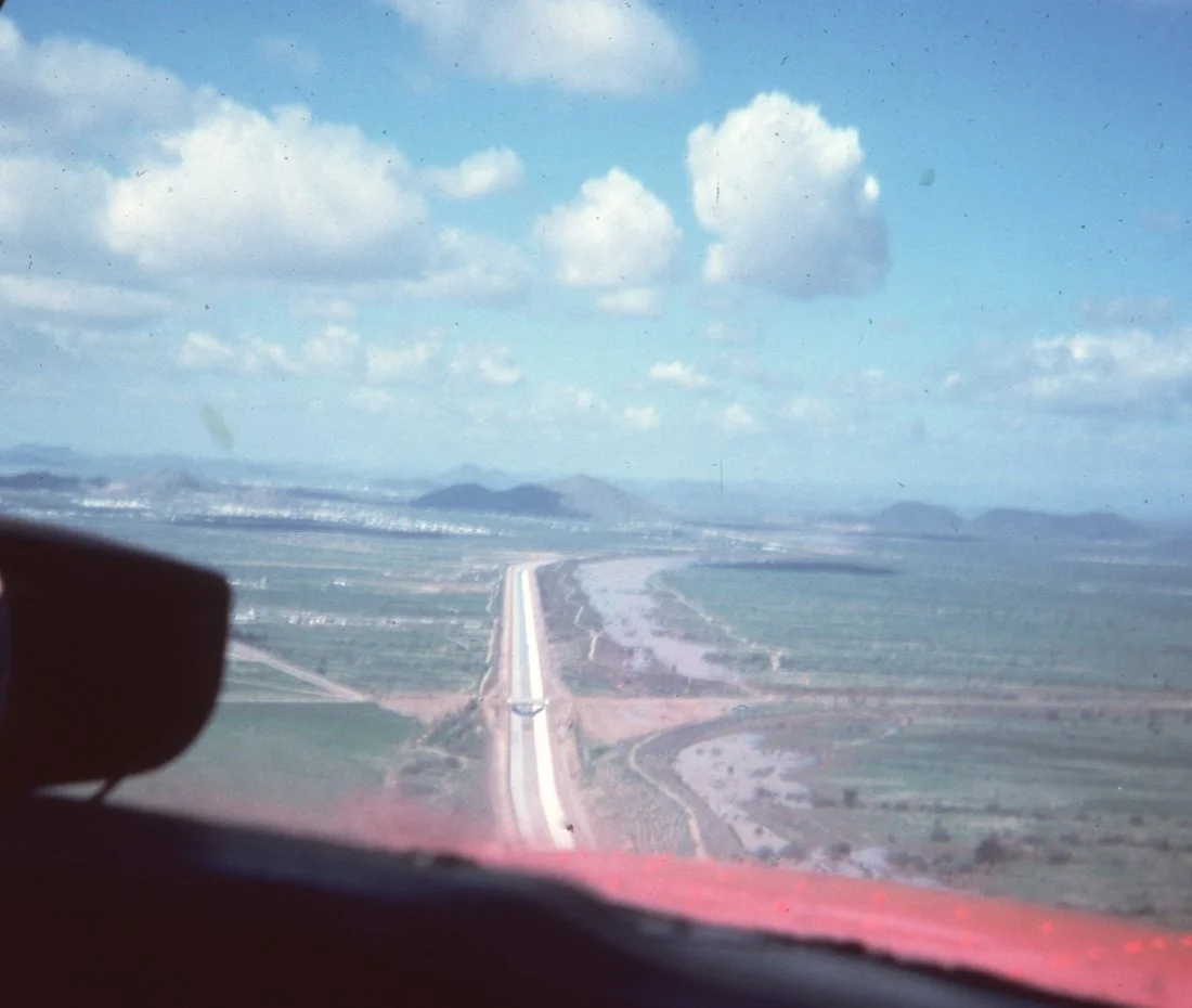 Aerial view from an airplane window showing a long road stretching across a flat, green landscape with a river or water body running parallel. The sky is blue with scattered clouds.