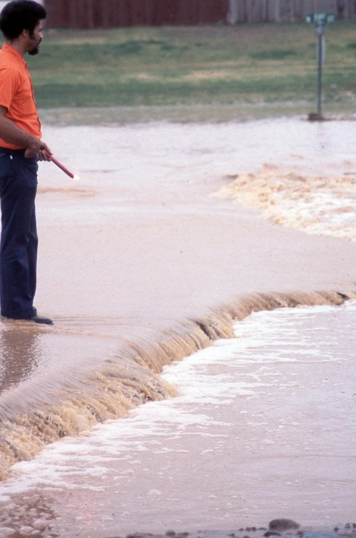 A man standing at the edge of a flooded area with muddy water, holding a red stick, with a partly submerged fence post in the background.
