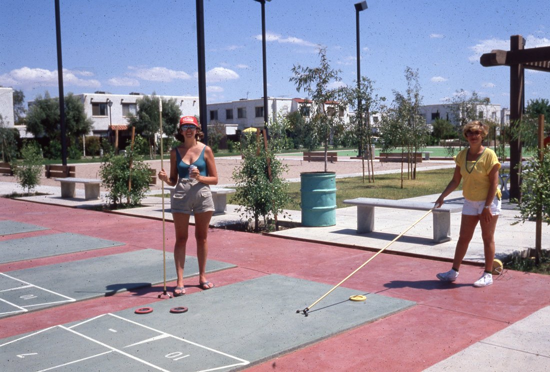 Two women are playing shuffleboard on an outdoor court with painted lanes and scoring areas, surrounded by small trees and benches, under a blue sky with some clouds.