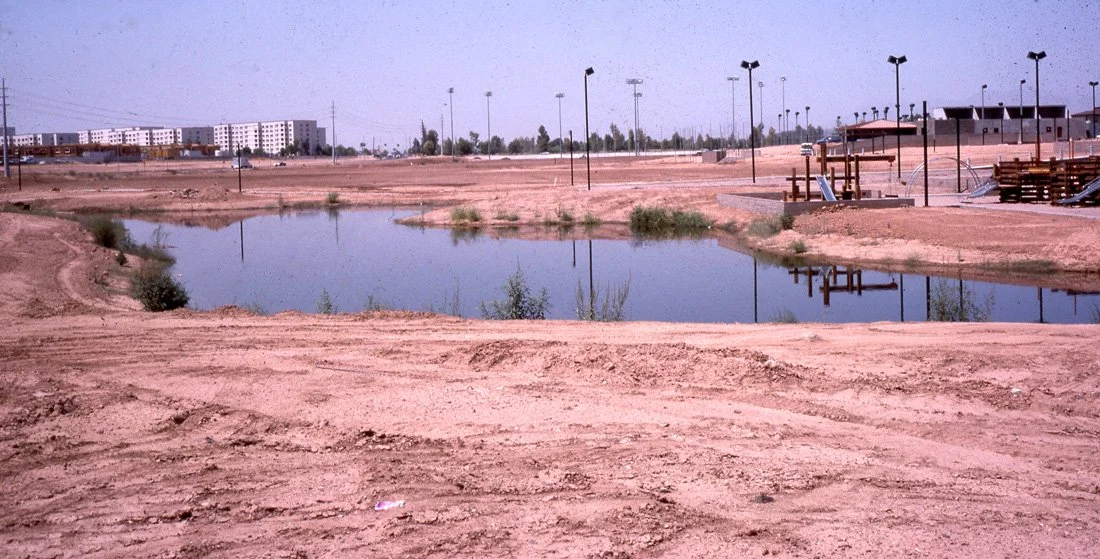 A dirt empty lot with a small water pond, some scattered bushes, a playground with slides and swings, and streetlights, with buildings in the background.