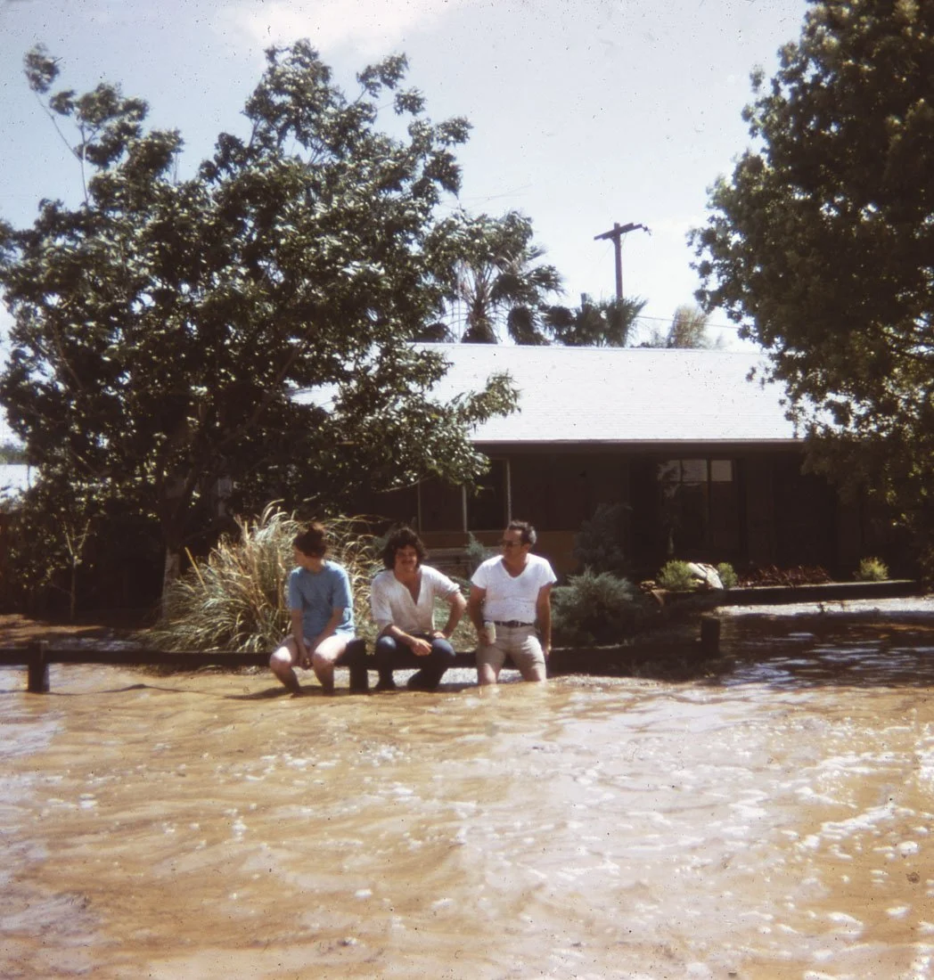 Three people sitting on a wooden and metal barrier in front of floodwaters near a house surrounded by trees and bushes.