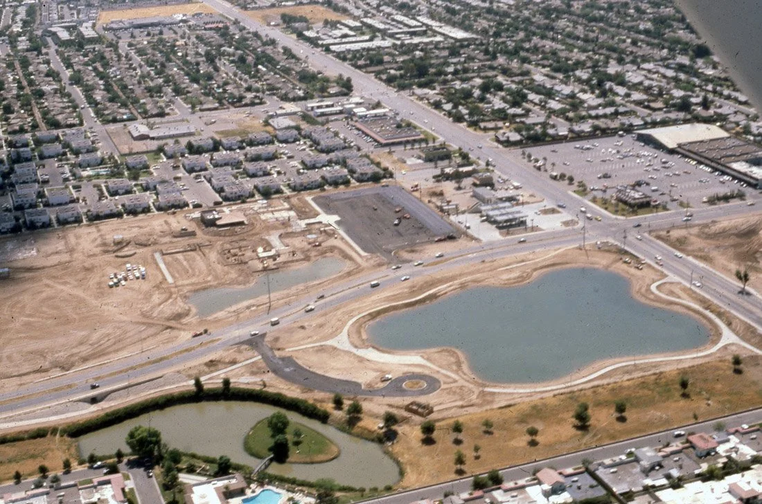 Aerial view of a developing area with a large pond, smaller pond, road, and nearby residential and commercial buildings.