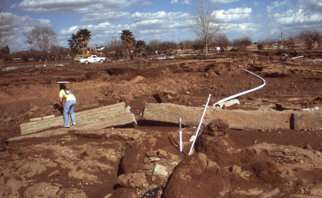 A woman working on a construction site with dug-up earth, pipes, and construction equipment in the background.