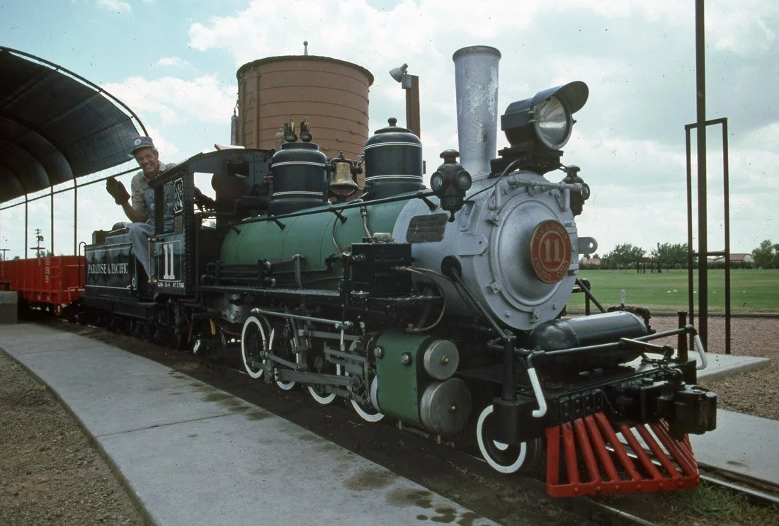 A vintage steam locomotive at a train station platform, with a person waving from the engine. The train is black with green and red accents.