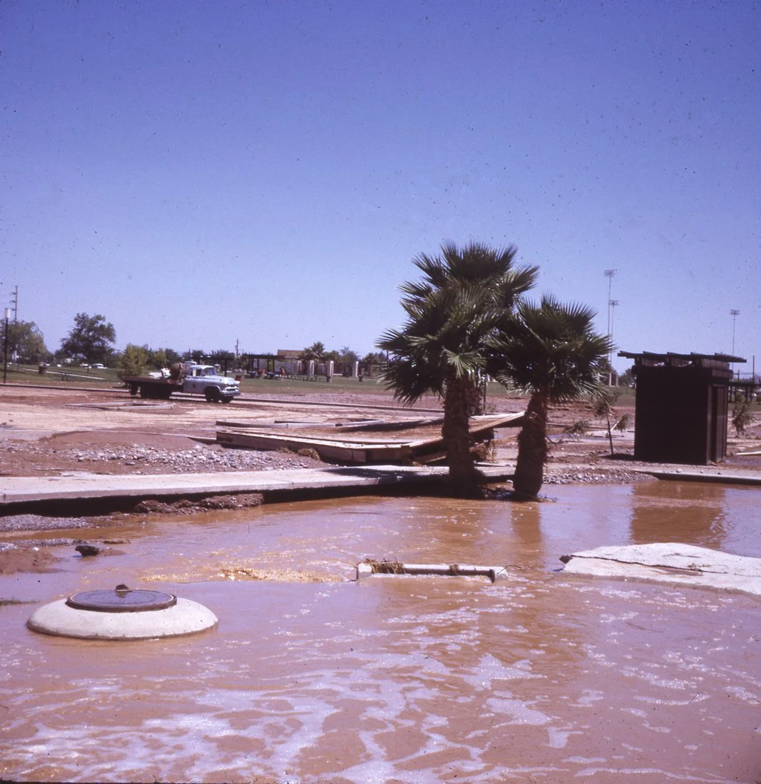 A flooded area with muddy water, two palm trees, and a small building. A pickup truck is parked in the background, and some structures are visible under a clear blue sky.