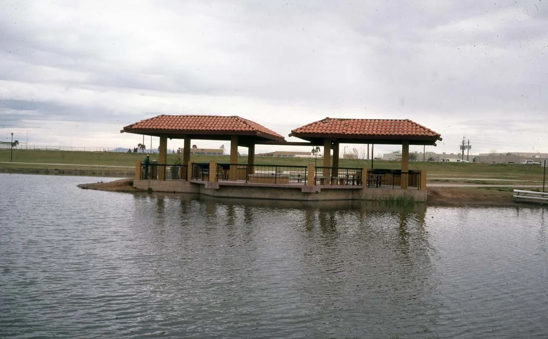 A covered pavilion over a body of water at a park with open grassy areas and cloudy sky in the background.