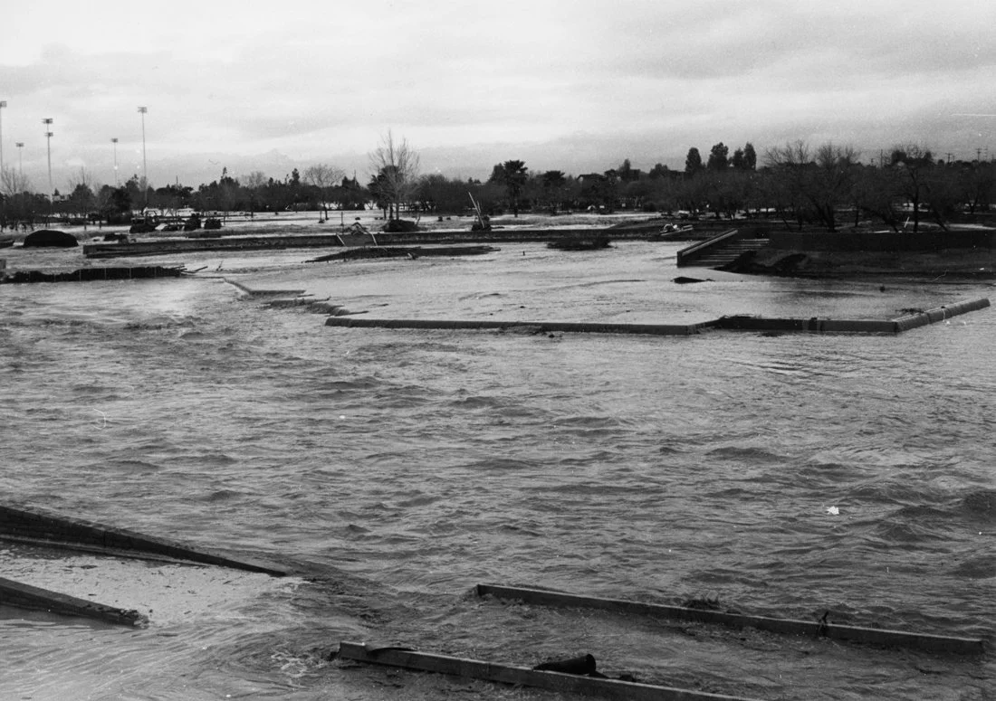 A flooded outdoor park with water covering pathways, trees, and structures under an overcast sky.