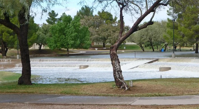 Park with a small waterfall or weir, trees, grass, paved walkway, and a water fountain.