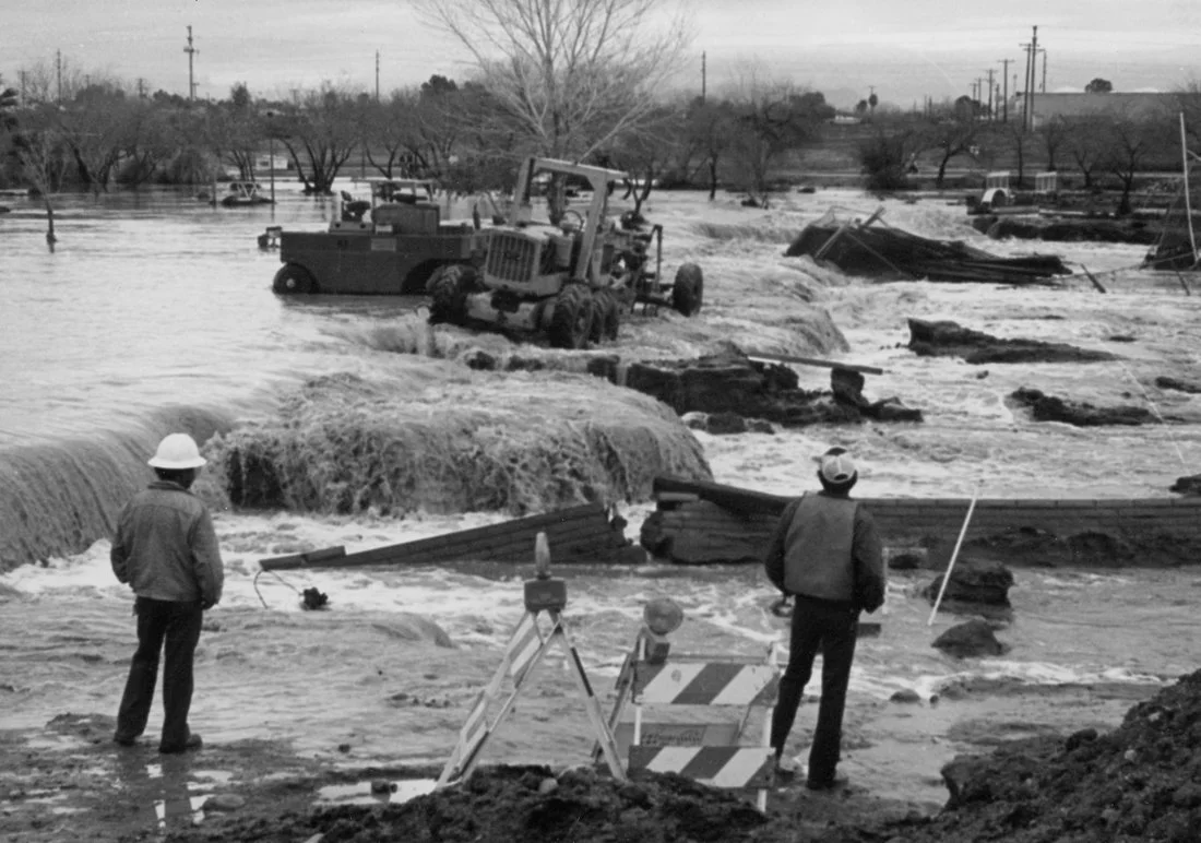Floodwaters submerge a flooded area with vehicles, construction equipment, and trees. Two men observe the scene, one with a hard hat, and rescue or construction signs are visible.