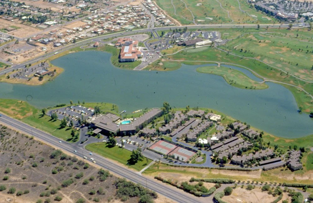 Aerial view of a large pond surrounded by residential, commercial, and recreational buildings, including a hotel, swimming pool, tennis courts, and golf course, near a highway in a suburban area.