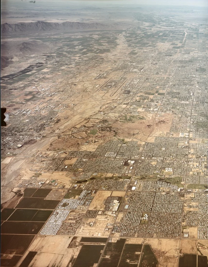 Aerial view of a desert city with grid-like streets, residential areas, commercial buildings, and agricultural plots, with mountains in the background.