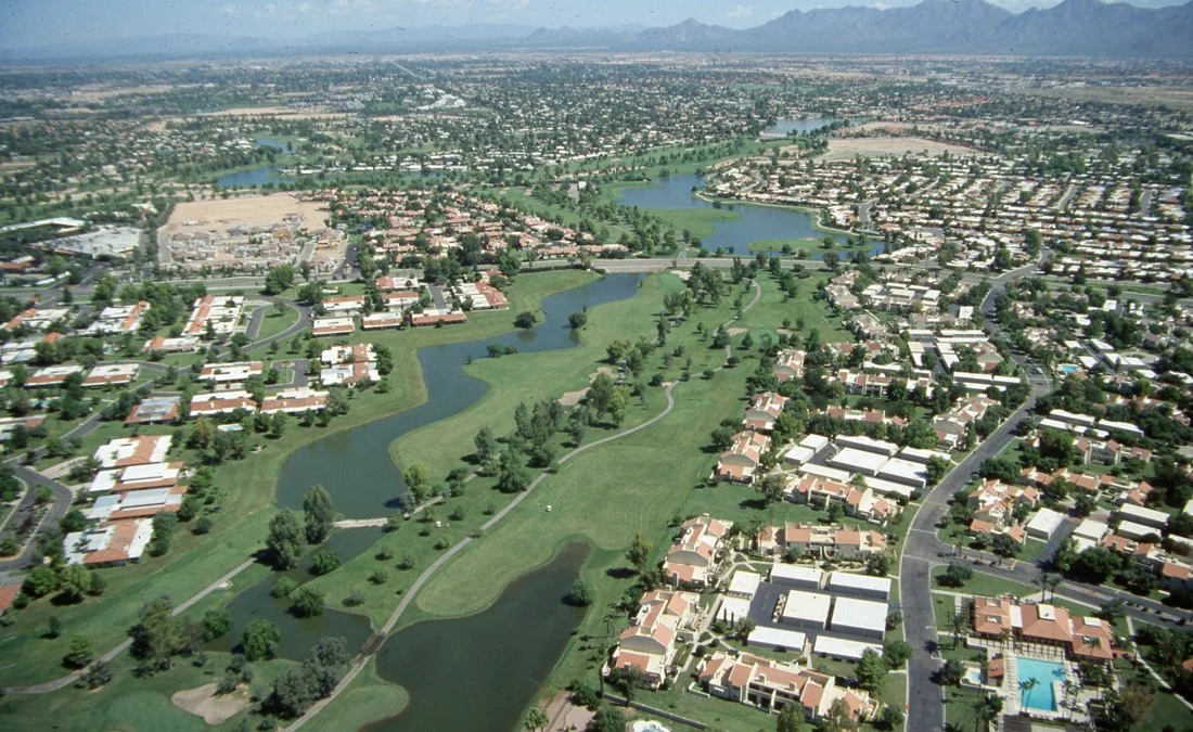 Aerial view of a suburban area featuring a golf course, lakes, and residential neighborhoods with houses and roads, mountains in the background.