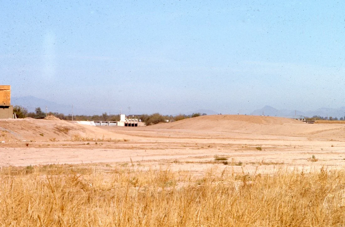 Desert landscape with dry grass in the foreground and sand dunes further back. There are some distant buildings and structures, with mountains on the horizon under a clear blue sky.