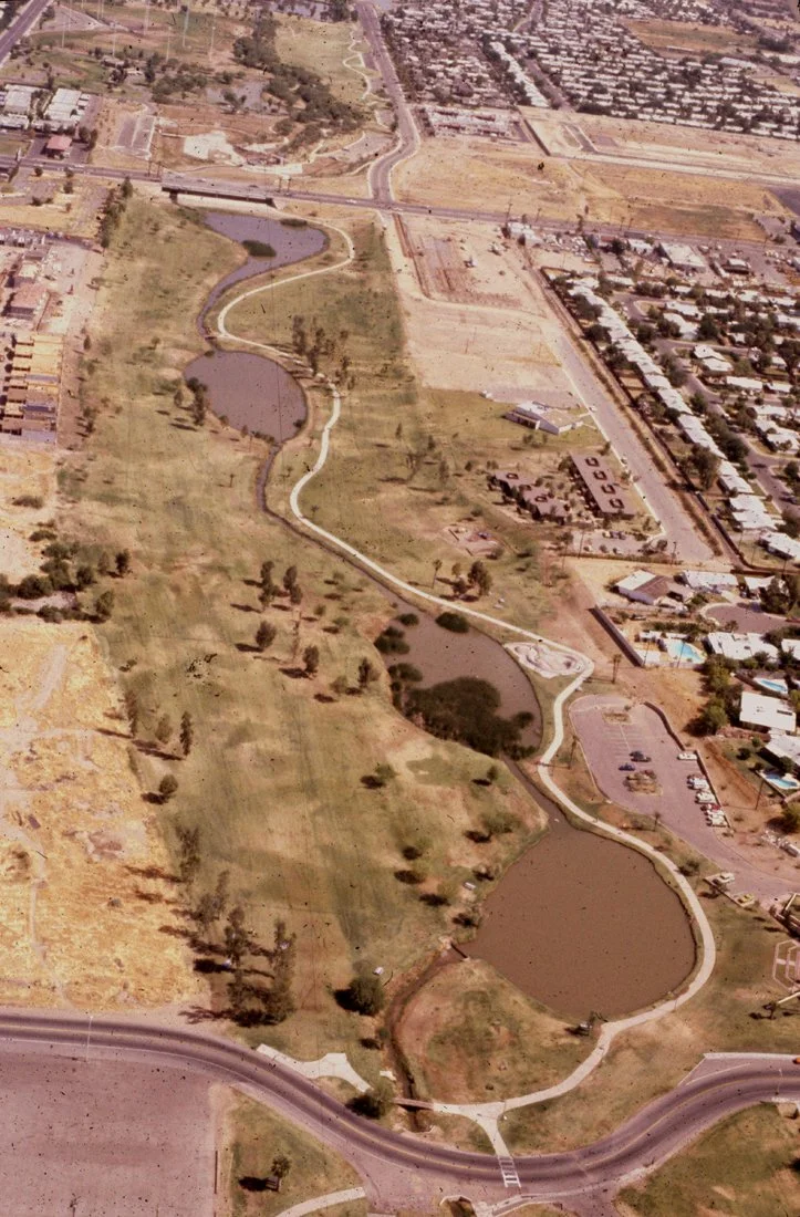 An aerial view of a park with a winding pathway and small lakes, surrounded by residential and commercial buildings.