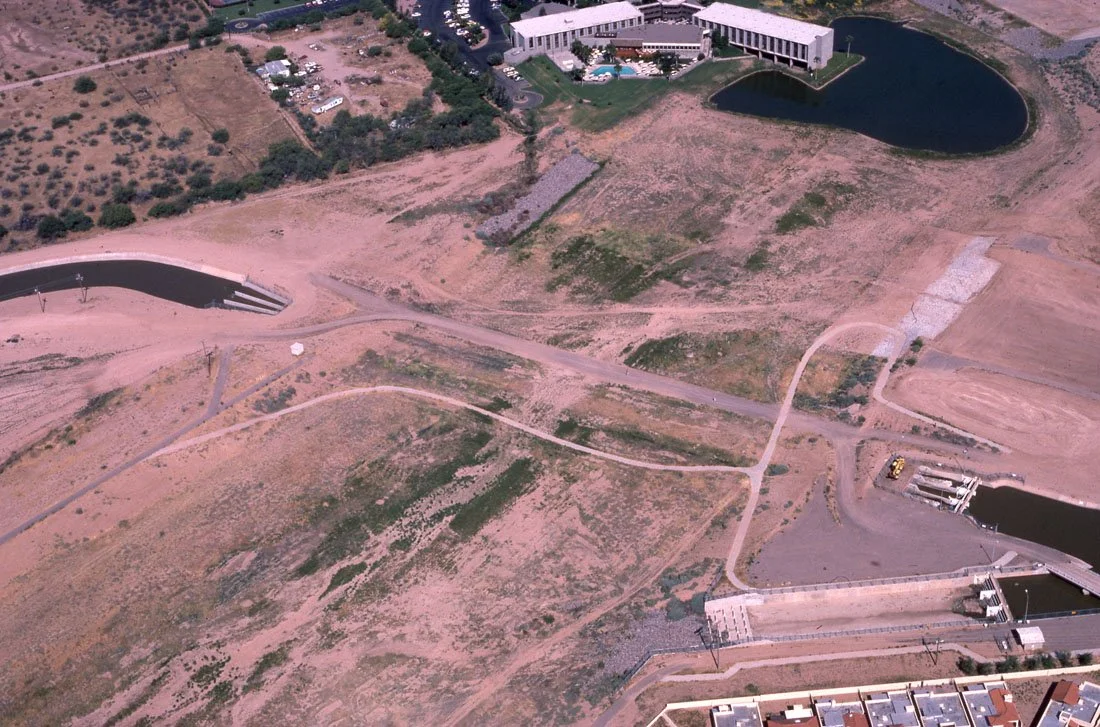 An aerial view of a large water reservoir with an adjacent modern building, open land with walking paths, a small pond, and some construction or industrial area nearby.