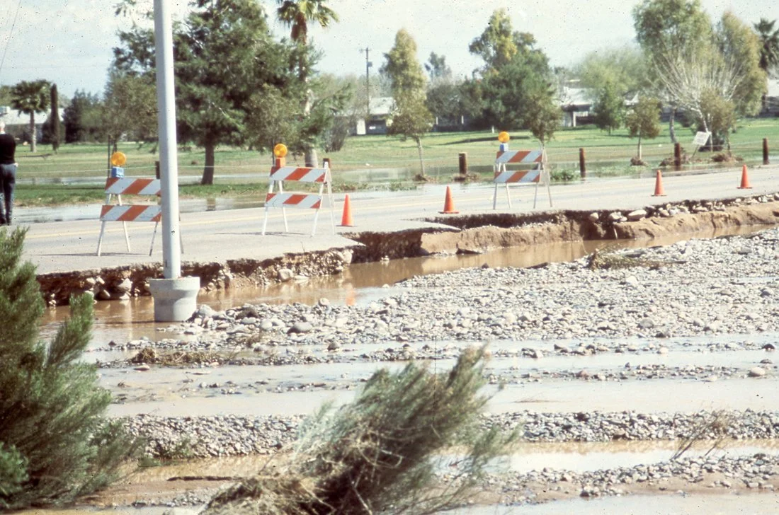 Road construction site with barriers, cones, a street light, and a large sewer or drainage ditch in the foreground.