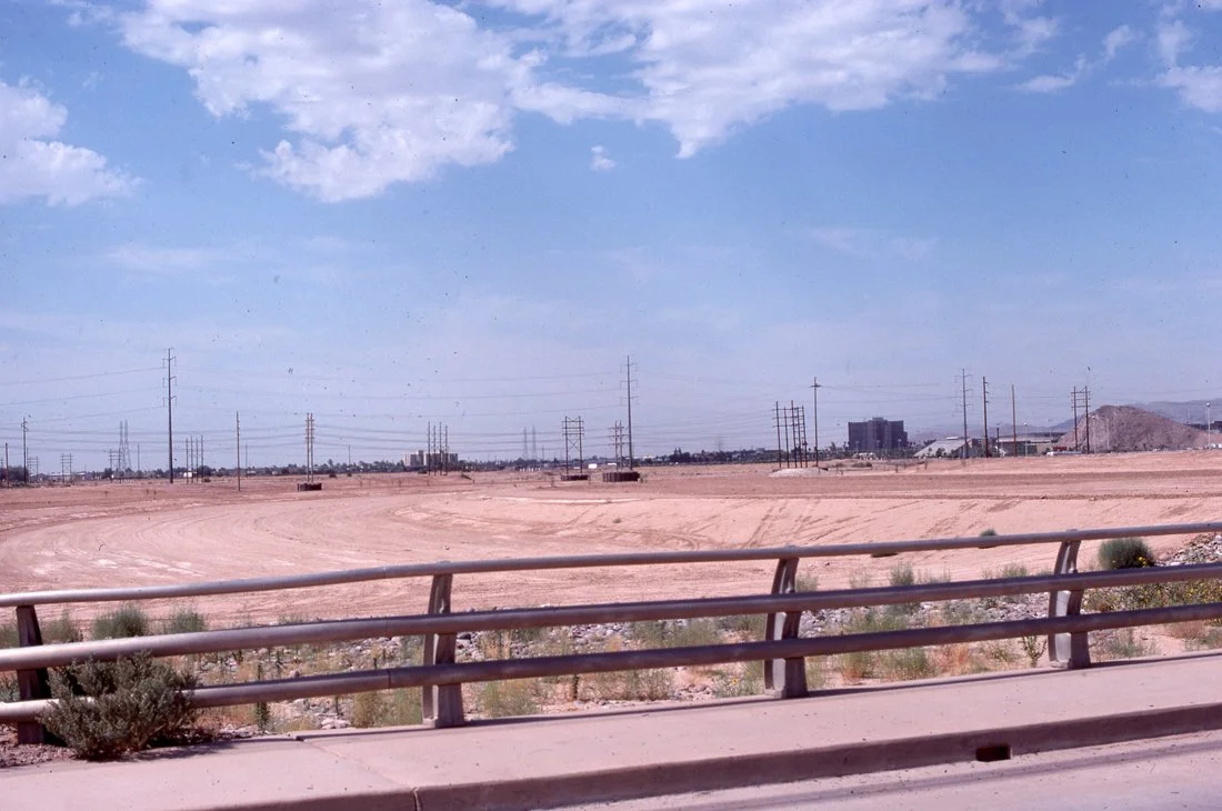 A barren desert landscape with power lines and distant buildings under a partly cloudy sky, viewed from a roadside with a metal guardrail.
