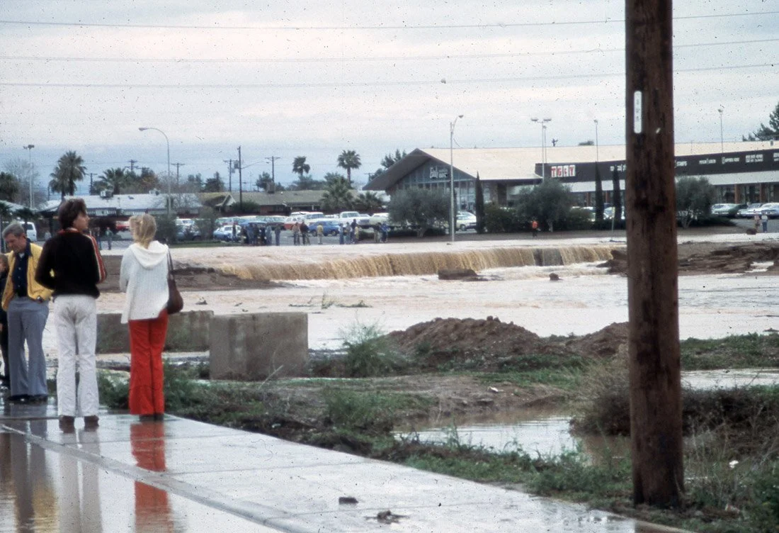 Street flooded from heavy rain with water overflowing onto the sidewalk, people standing under umbrellas in the background, and a power pole in the foreground.