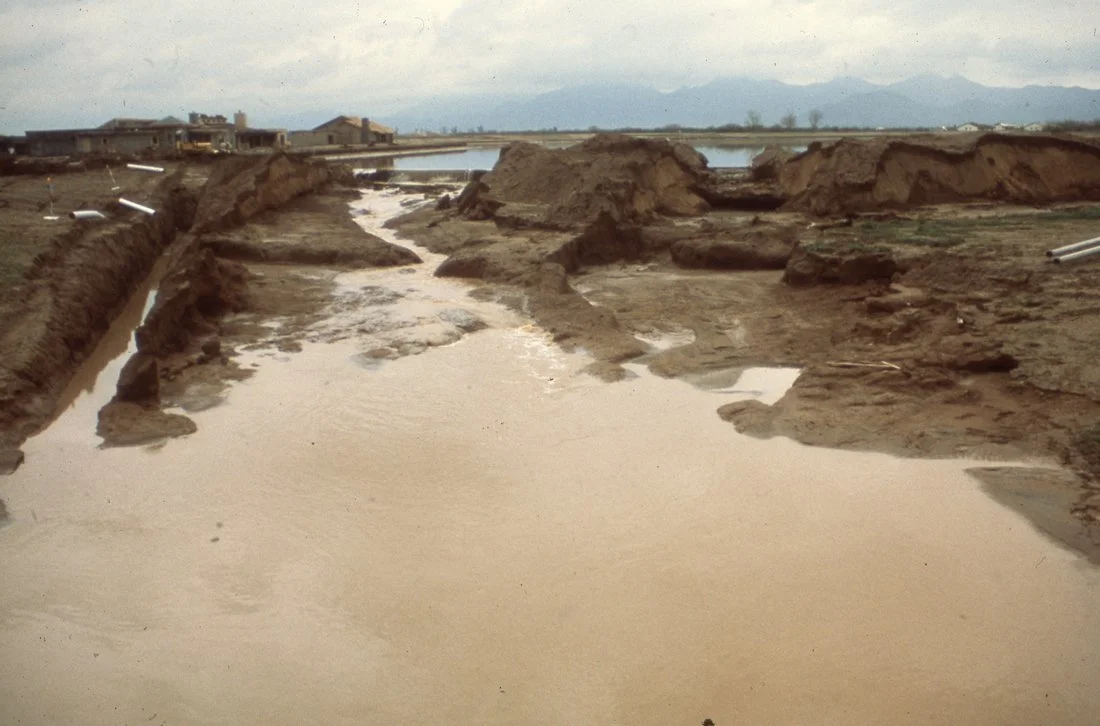 Construction site with earth excavation, trenches, and water runoff leading to a body of water in the background, with buildings and mountains.