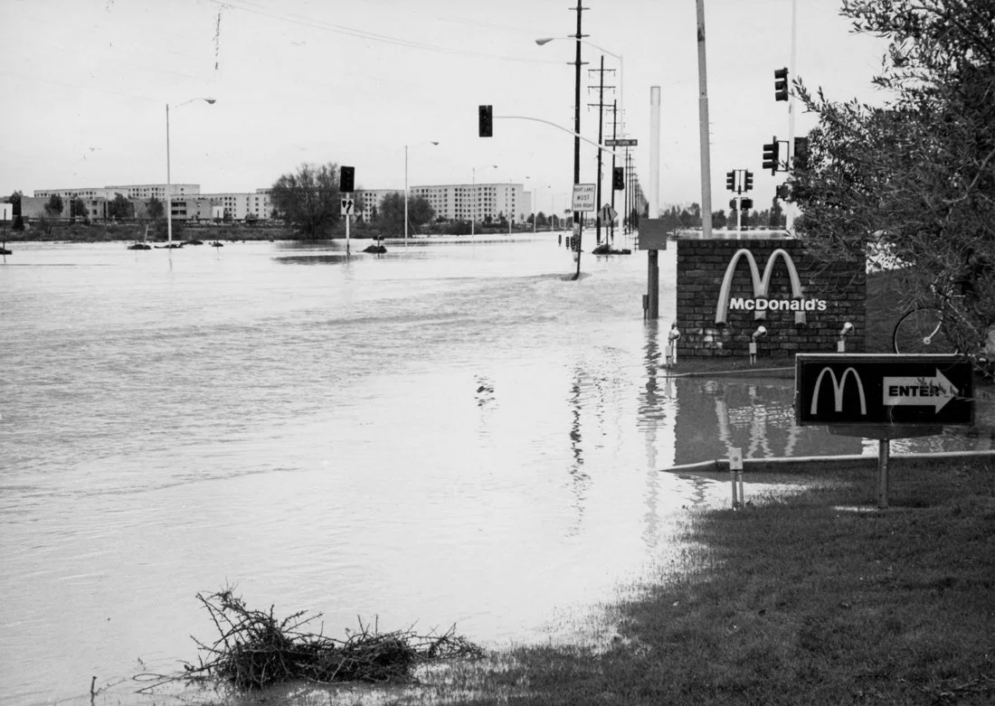 Flooded parking lot with water covering the ground, submerged vehicles in the distance, and a McDonald's sign on a brick wall with an entrance sign pointing right, amidst trees and power lines.